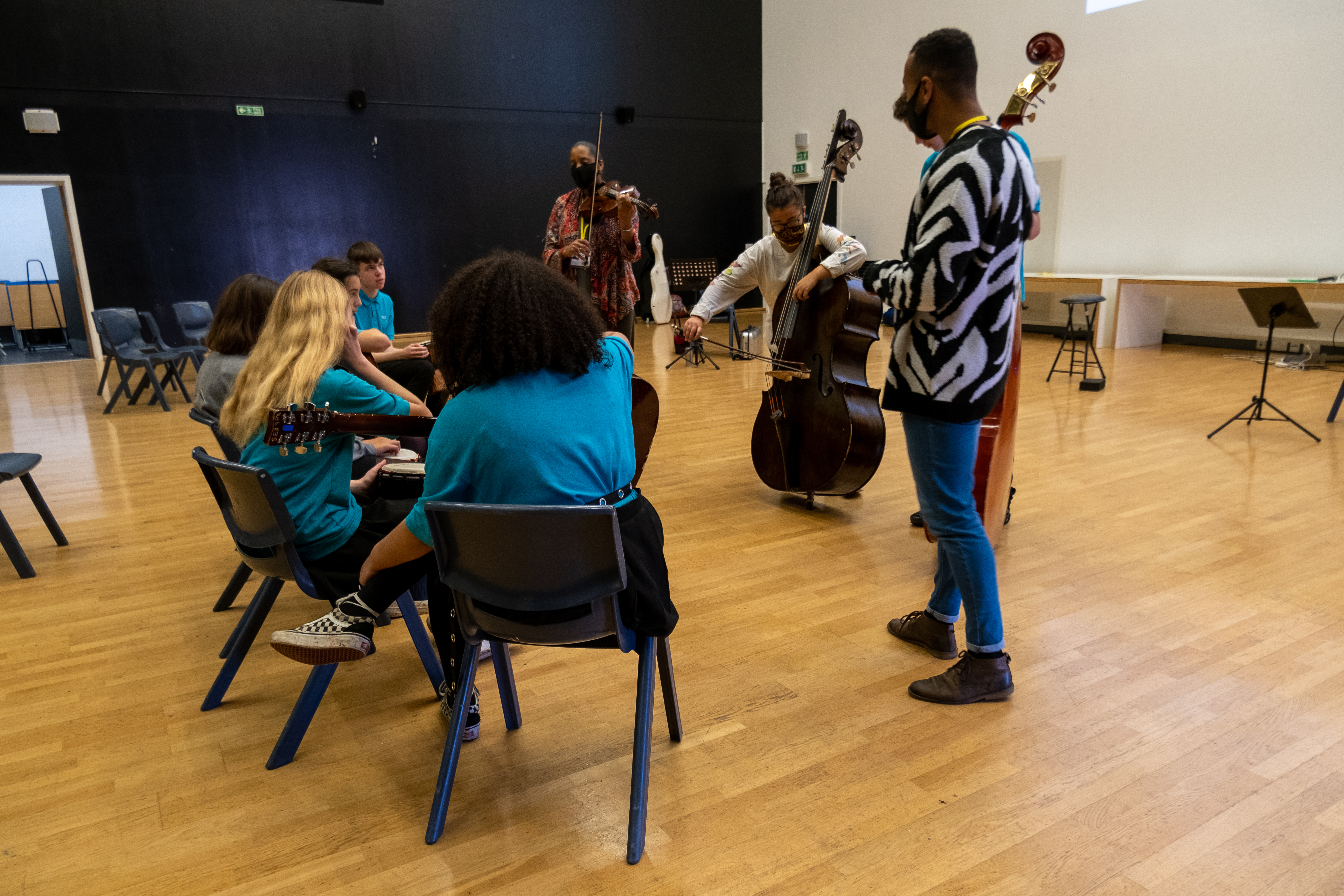 Musicians with instruments standing up to deliver workshops to school pupils sitting on chairs.