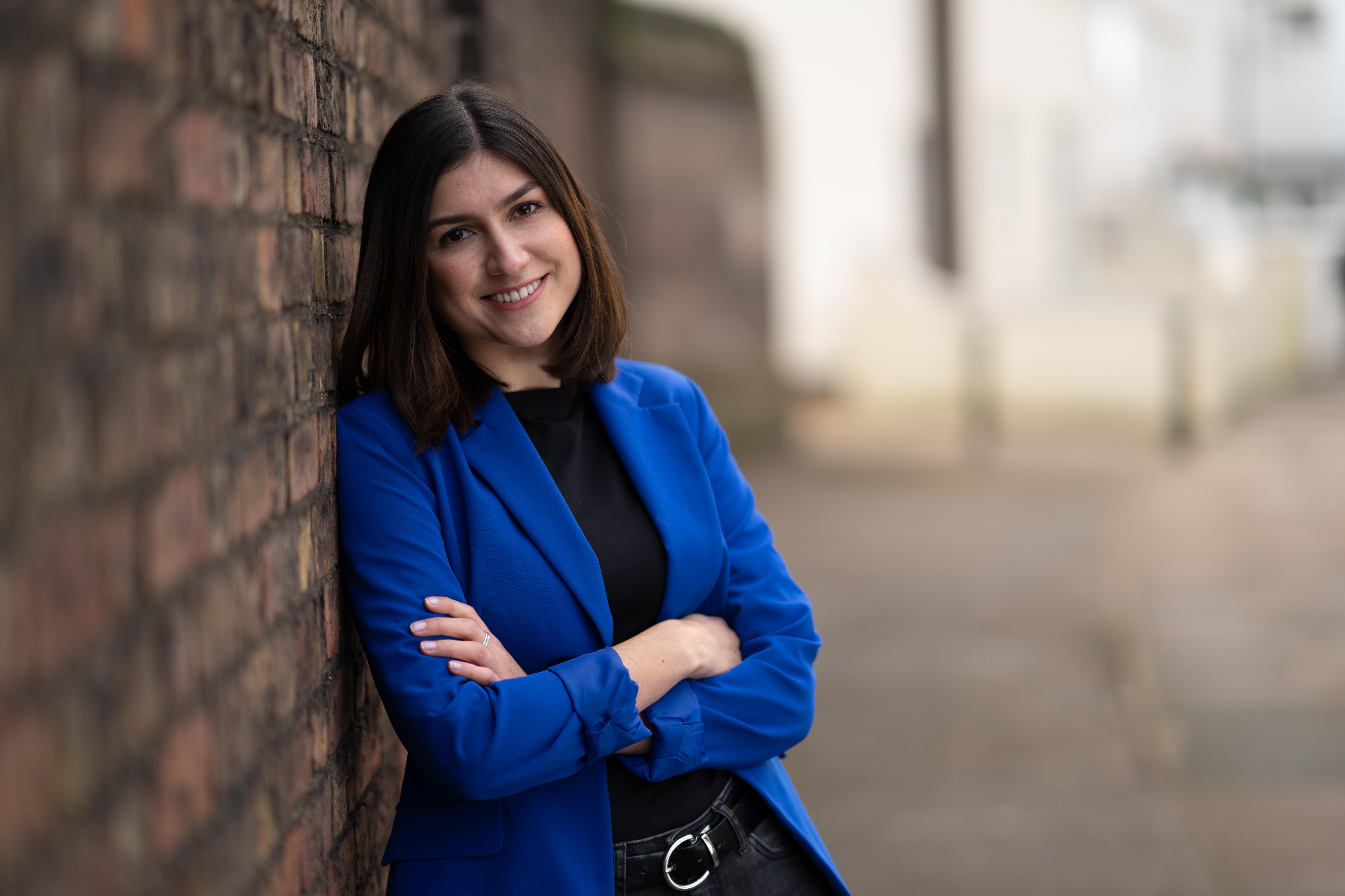 Person in blue coat with long brown hair leans against a brick wall whilst smiling and crossing their arms