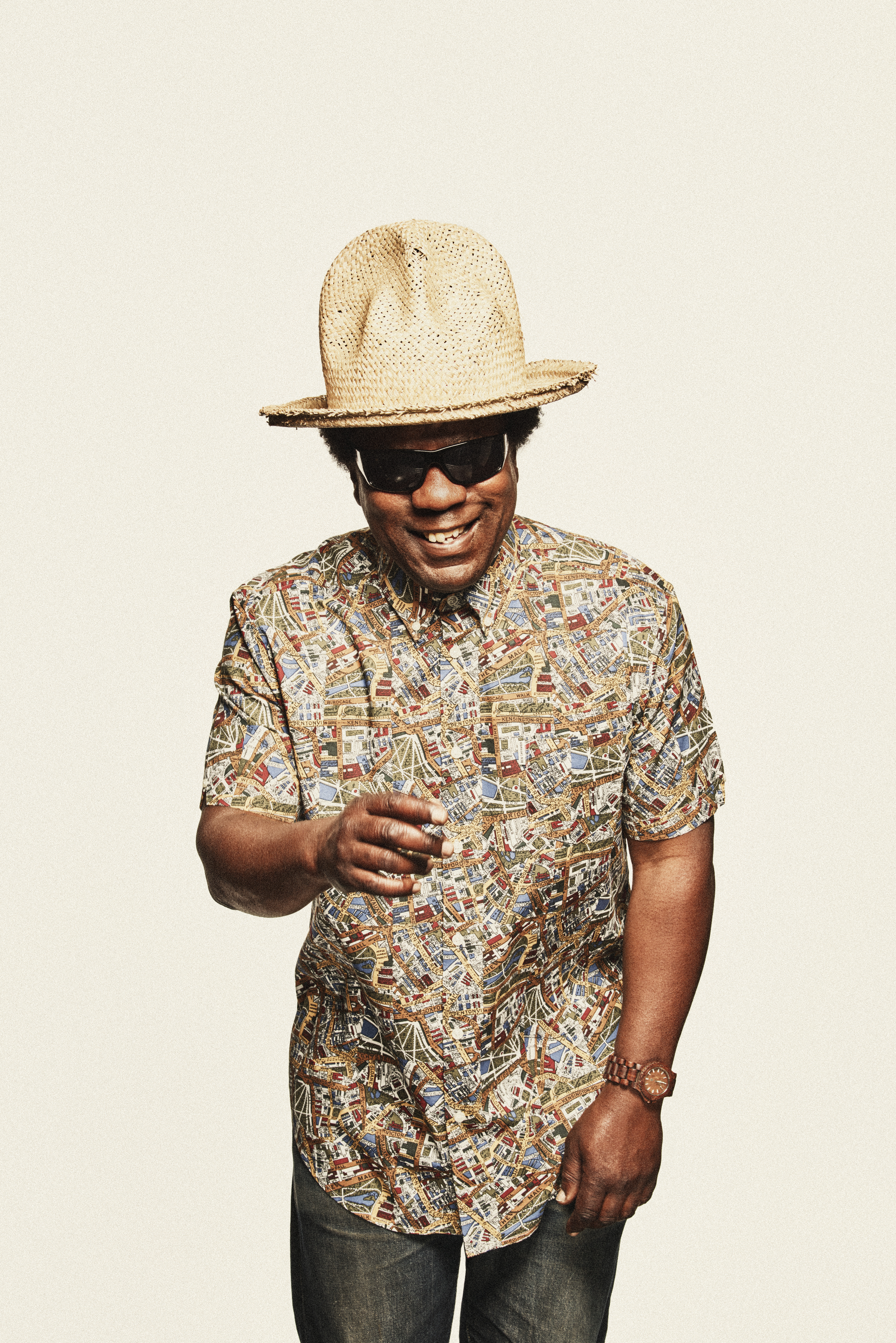 DJ Norman Jay smiles and points at the camera. He is wearing a cream bucket hat and a multi-coloured shirt.