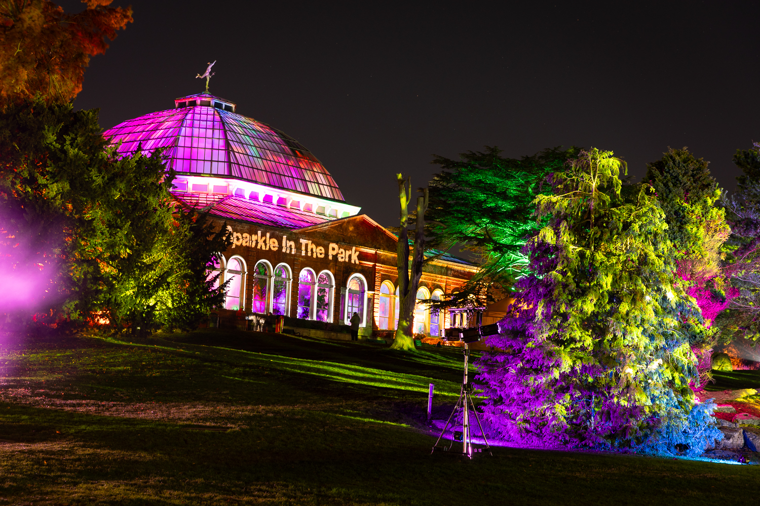 A large glass building (Avery Hill Winter Garden) lit up in rainbow colours. The words 'Sparkle in the Park' are projected on to it in white.