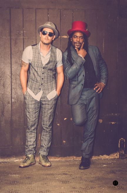 Tyber and Pete from The Dualers. Two men in hats leaning up against a wall. Both wearing casual suits. The man on the left wears a grey outfit and the man on the right wears blue. 