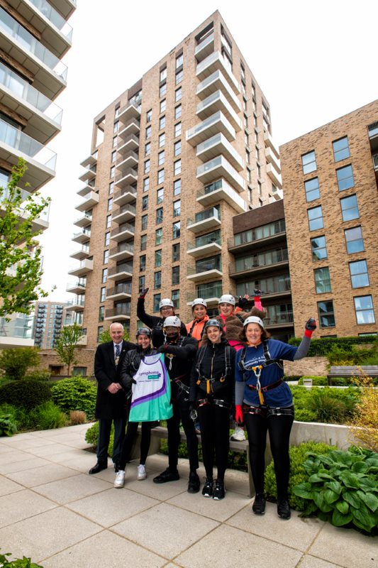 The mayor stands in front of  17 storey building with seven people dressed in abseiling equipment. They are cheering and holding up a vest top with 'Lymphoma Action' printed on it