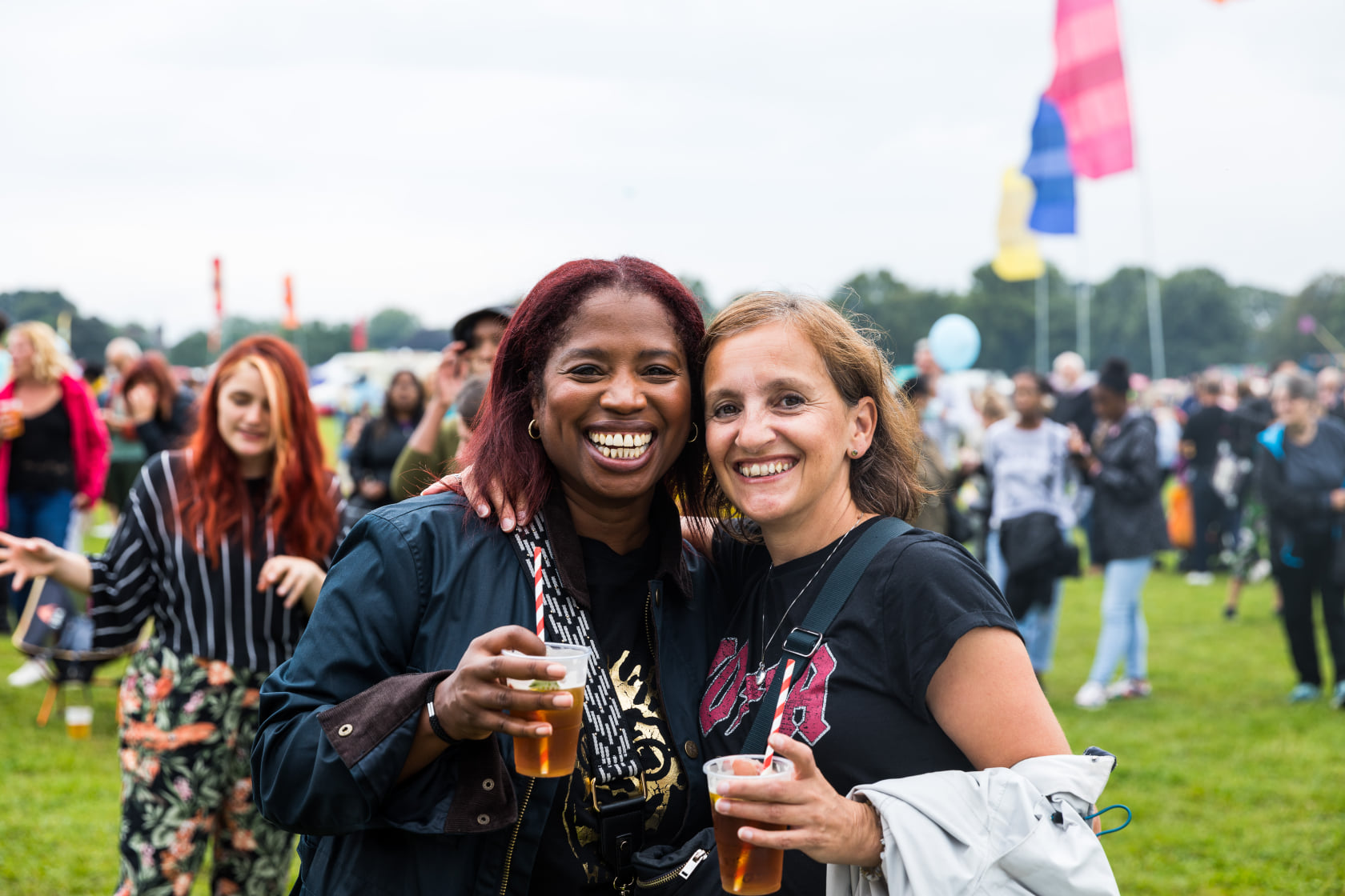 Two women stand smiling at the camera holding drinks at a festival. There are people walking past out of focus in the background. 