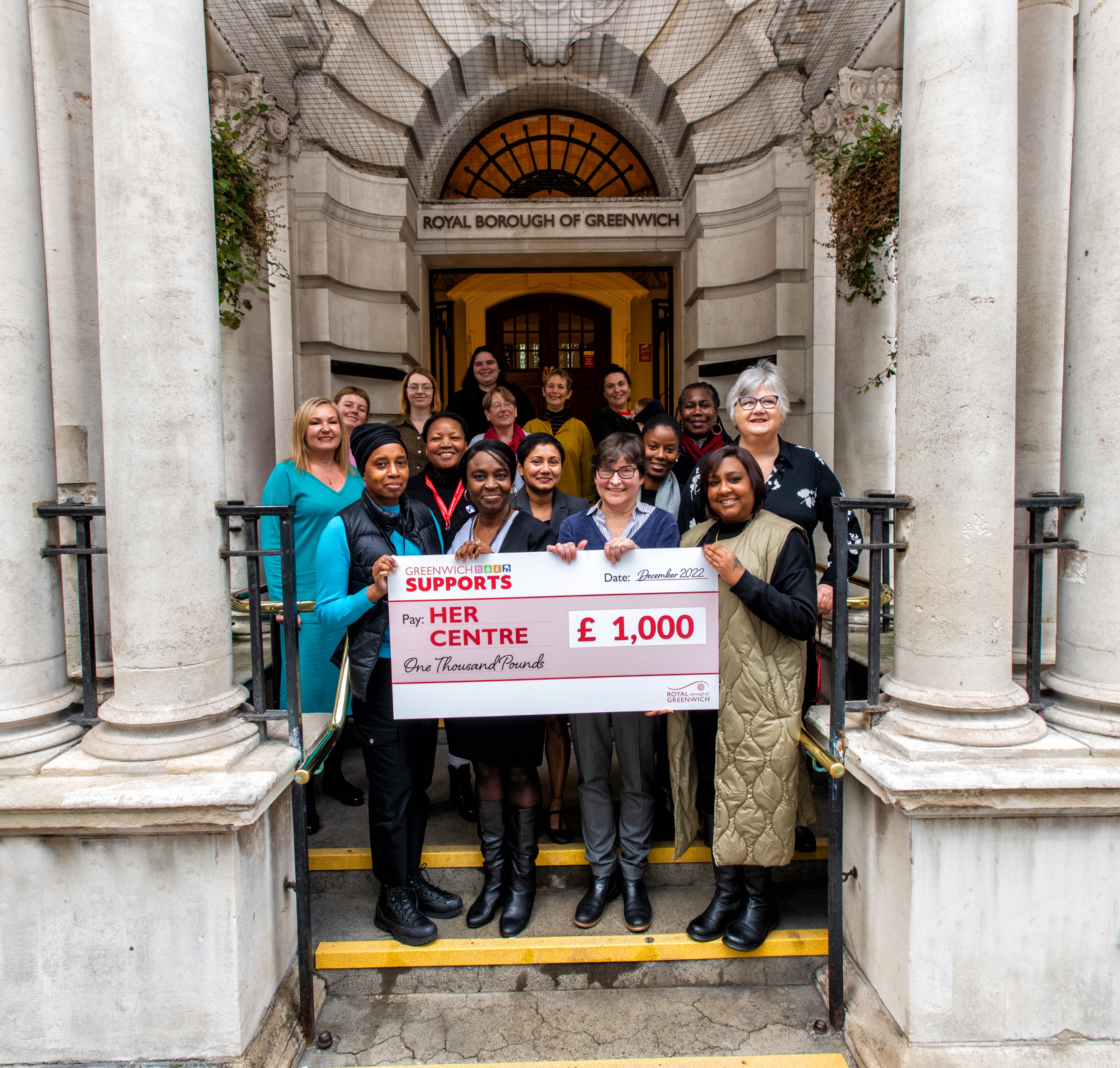 Picture of a group of women from the Council and HER Centre outside the steps of the Woolwich Town Hall, holding a large cheque for £1,000