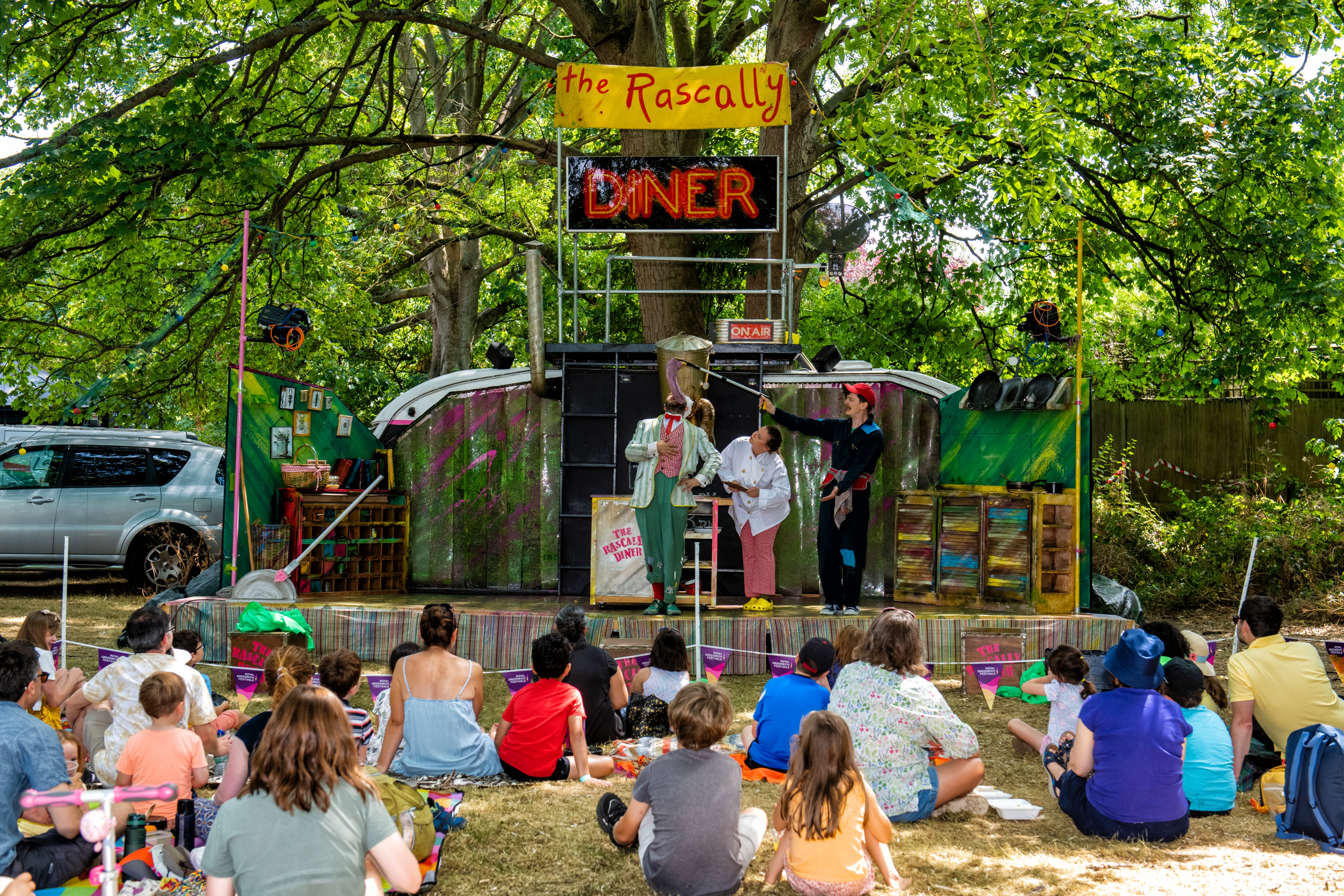 A group of children face a stage watching a two performers on stage. A sign above the stage reads 'Rascally Diner'