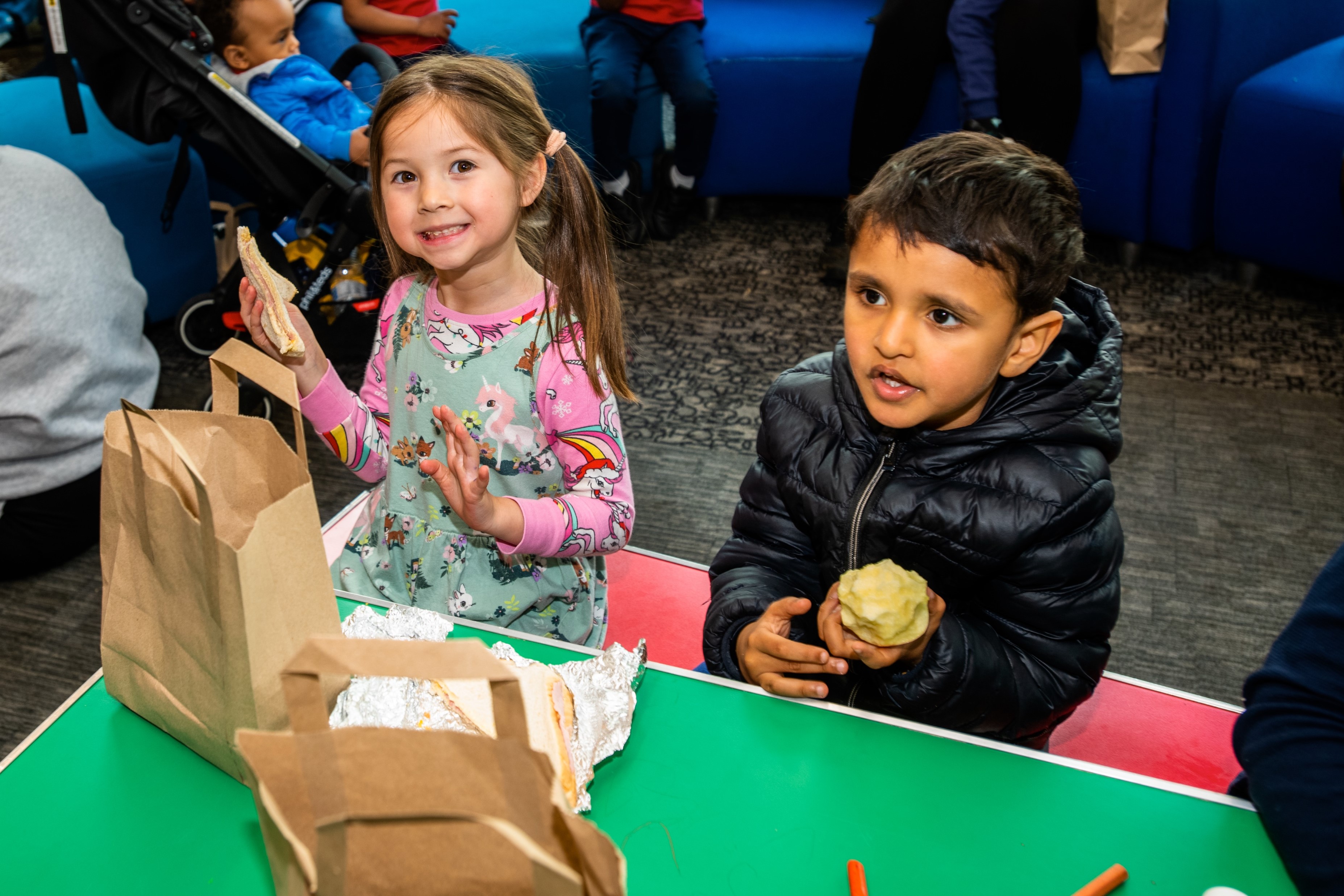 A young girl and boy enjoying a packed lunch. The girl holds and sandwich and a boy holds an apple.