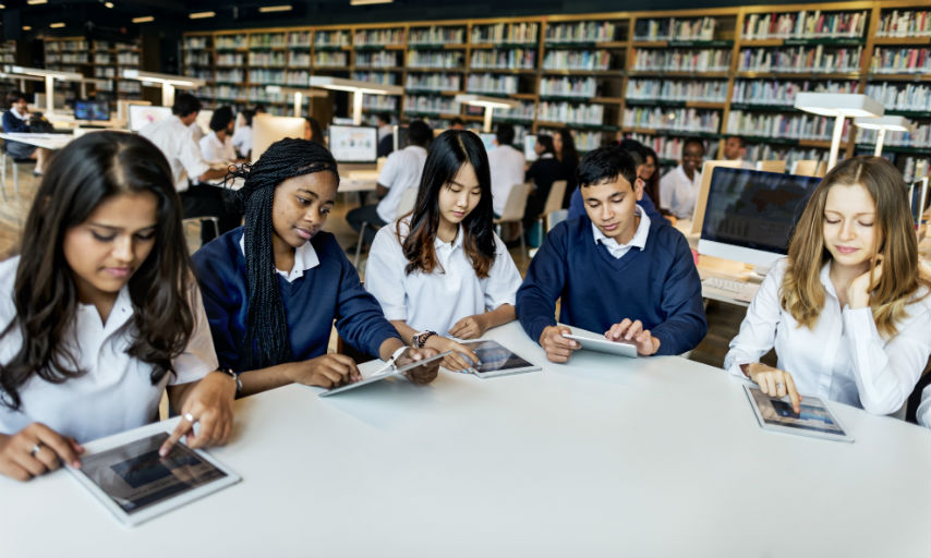 Five school students in a school library, sat around a table. Each student is holding a tablet in their hands.