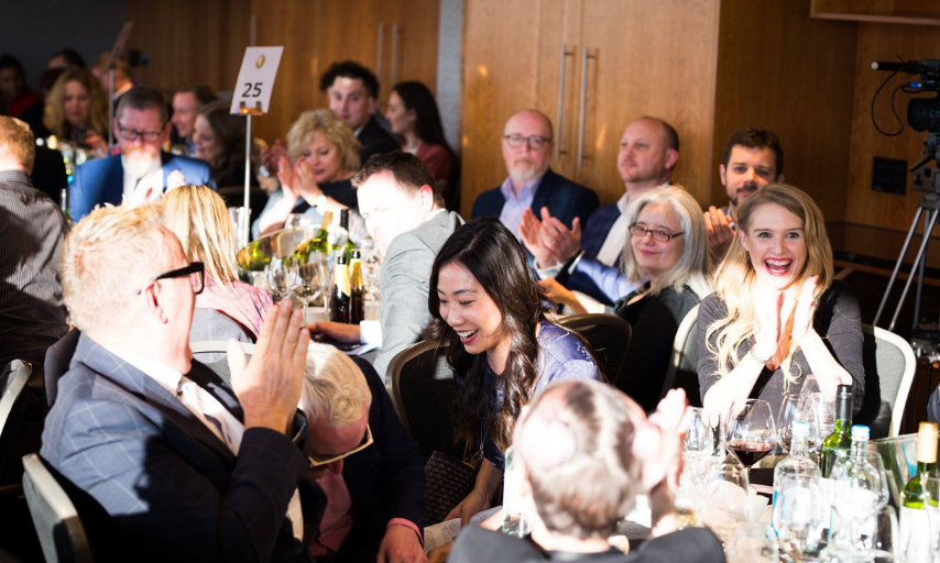 People sit across different tables at the Business Awards. Many are clapping and looking towards a woman with dark, wavy hair who is centre of the photo smiling and looking to the left.