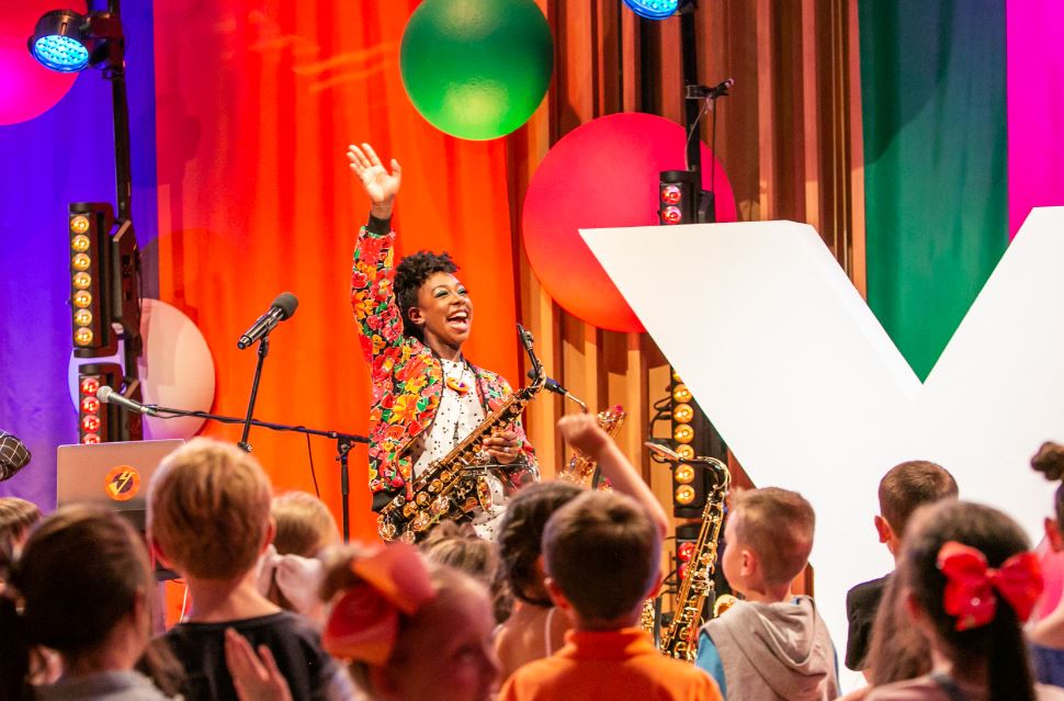 YolanDa's Bans Jam performing on a colourful stage. She holds a saxophone as she waves to her audience of smiling children.