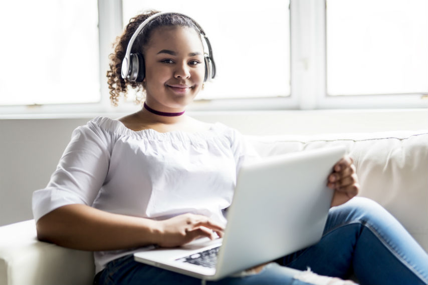 Smiling teenage girl with silver headphones on, sitting on a cream sofa, holding a white laptop.