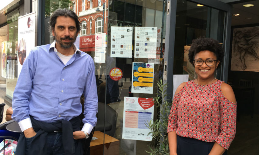 A white man in a blue shirt standing next to a smiling black woman who's wearing black glasses and a red blouse. They are both standing outside the artFix cafe in Woolwich.