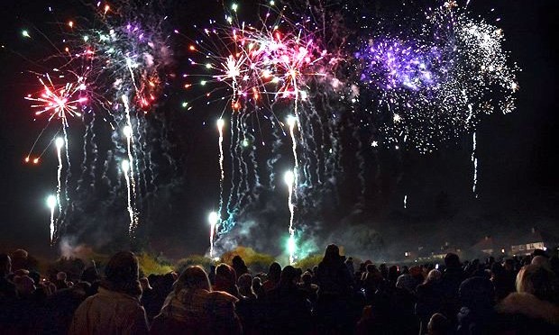 Fireworks display of red, white, purple and green bright light against the dark night's sky, with the silhouettes of a large group of people observing the fireworks displayed above.