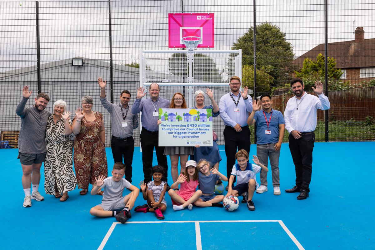 A group consisting of councillors, council staff, centre users and contractors waving in the MUGUA at Middle Park Community Centre logo. The writing on the board reads: "We're investing £430m to improve council homes - our biggest investment for a generation."