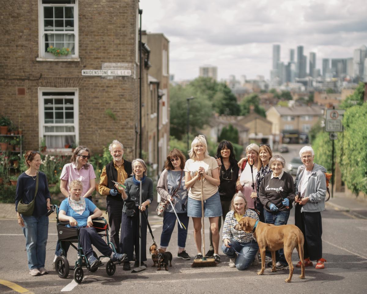 A group of people standing in the road smiling. There are also two sausage dogs and a big brown dog, and the people are holding litter picking and weeding equipment.