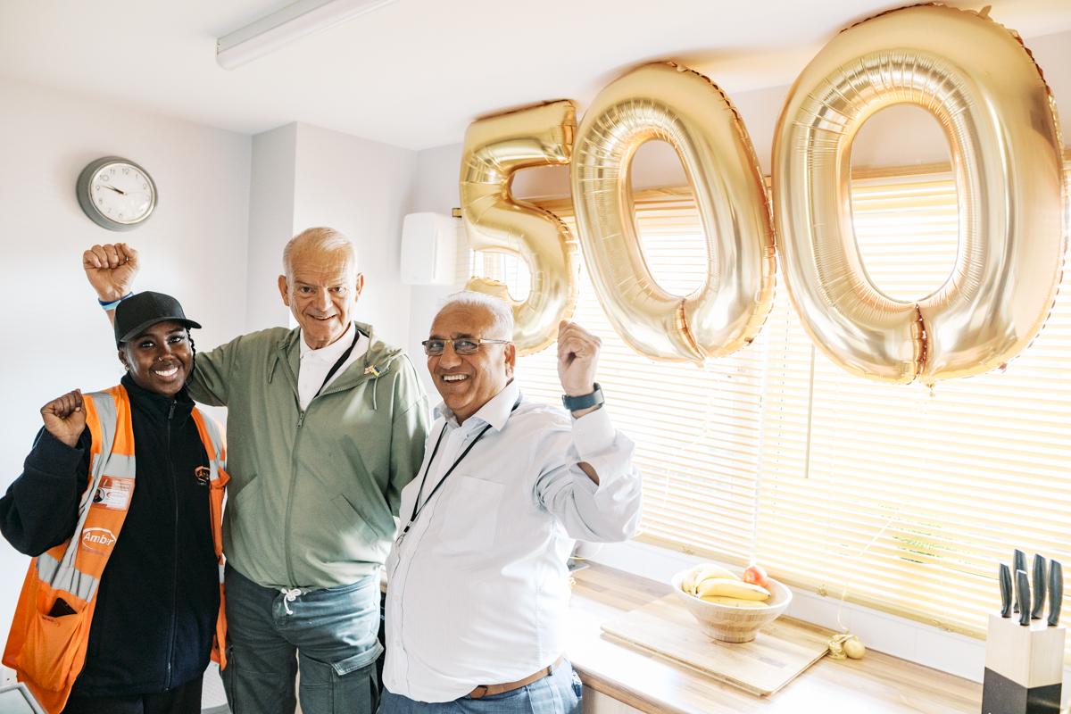 Three people standing in a kitchen with gold balloons displaying 5-0-0