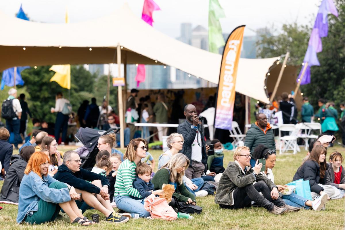 A group of people sitting down on grass at an outdoors event. Behind them is an open air tent and a flag that says 'tramshed'