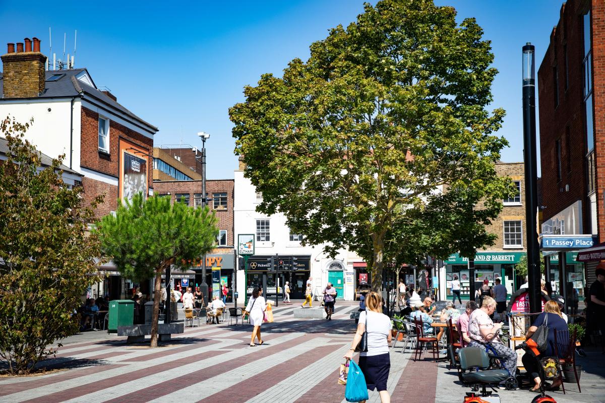 The square in Eltham High Street, people sit outside a cafe, people are walking around with shopping bags.
