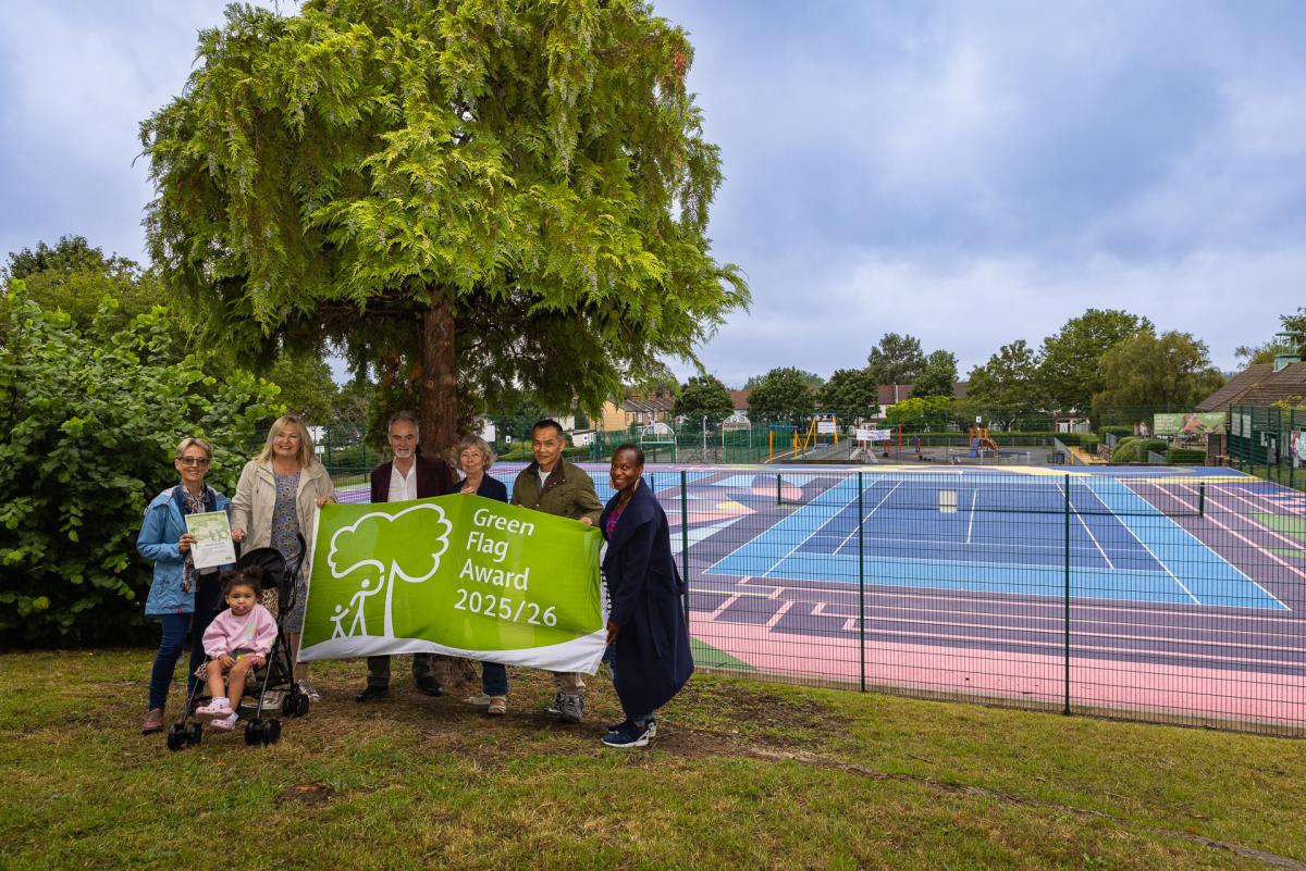 A group of people standing on grass holding a flag that reads 'Green Flag 25/26' A big tree is behind them and a tennis court beyond that.