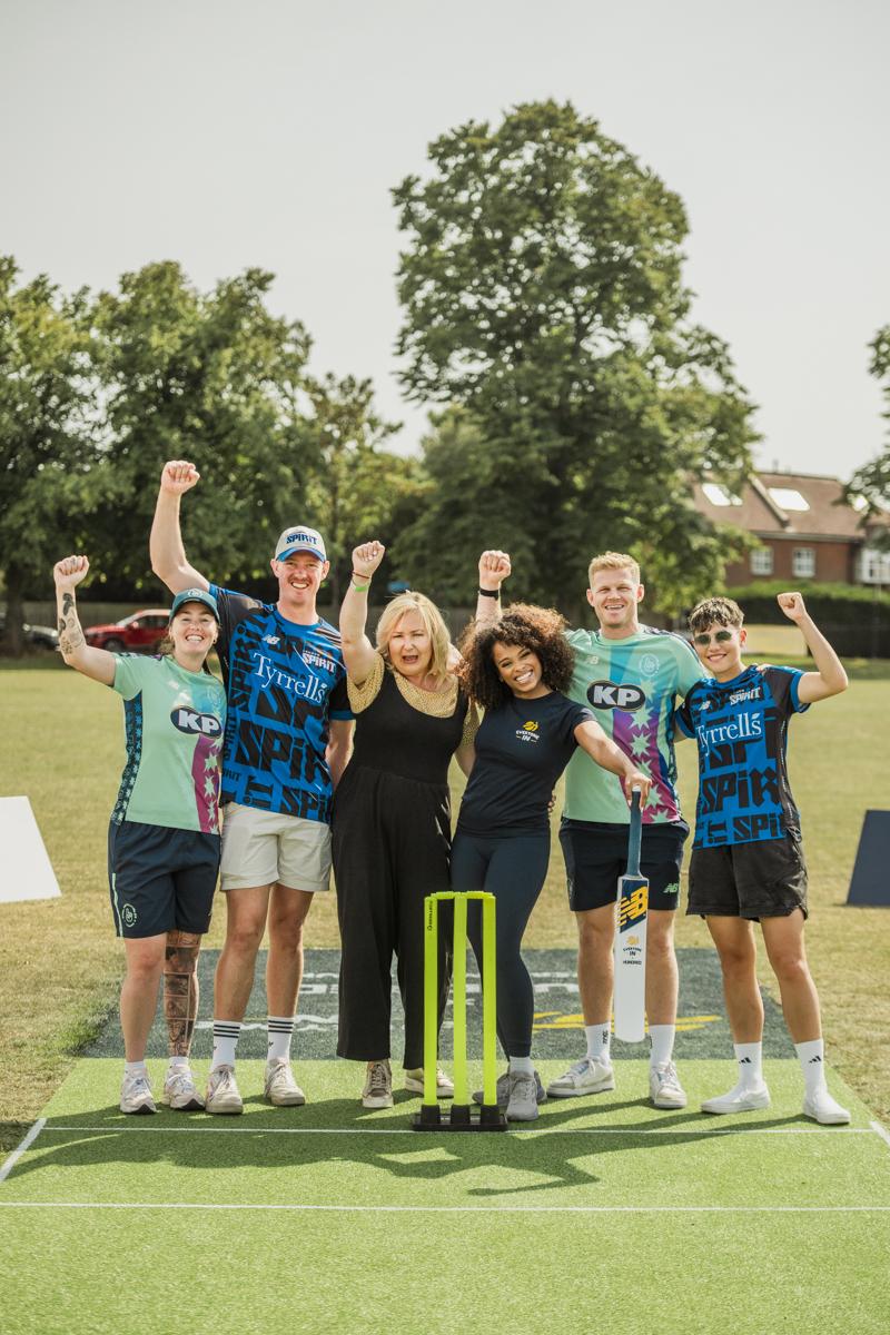A group of five people standing on a green artificial cricket wicket with their hands up in the air to cheer