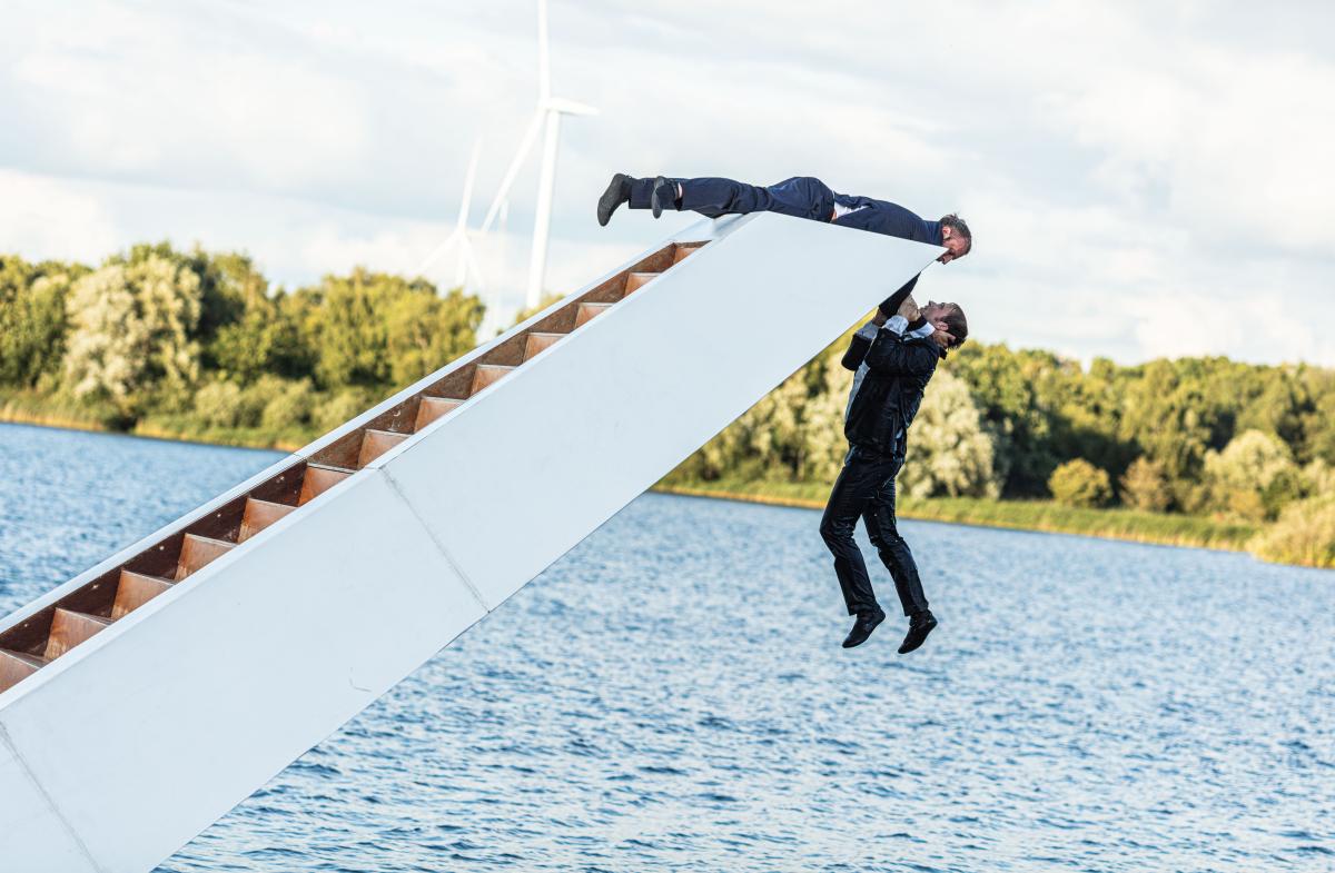 Two people hanging from a white structure floating on a lake, with green trees in the background