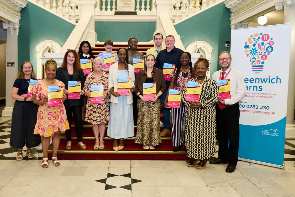 Group of people on a staircase holding certificates at a this year's Adult and Community Learning Awards at the Town Hall. To the right is a banner with a light bulb design that reads GREENWICH LEARNS.