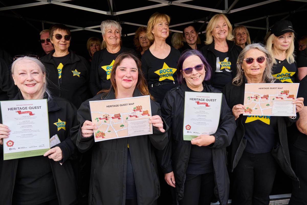 A group of people, some holding HERITAGE QUEST clues and HERITAGE QUEST MAPS, pose outdoors. They are wearing black shirts with yellow stars that read ROCK CHOIR. 