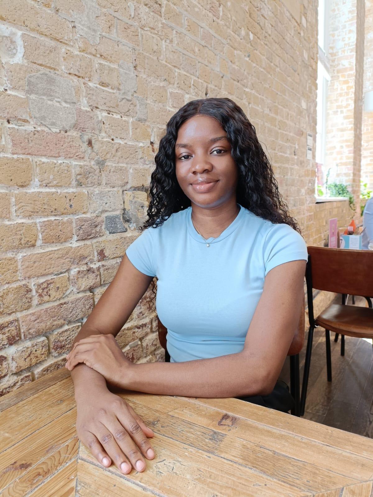 Blessing has long curly hair and is wearing a light blue shirt. She is sat at a wooden table in front of a brick wall, with hands resting on the table in a relaxed indoor setting. She is smiling and looking to camera.