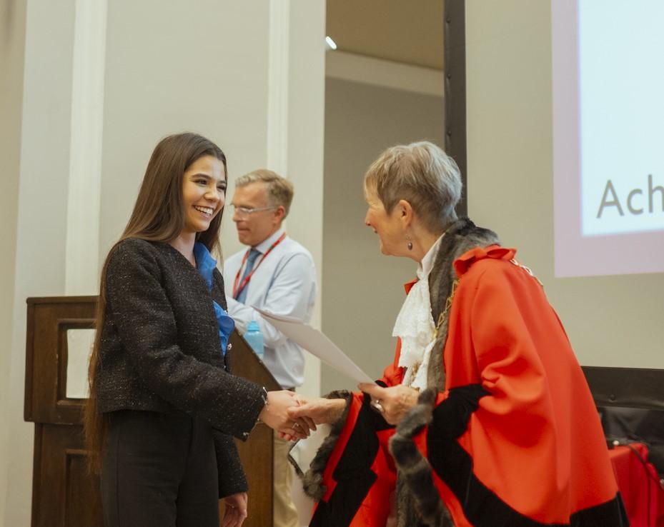 The mayor, in red mayoral gown, shakes hands with a young person who is smiling