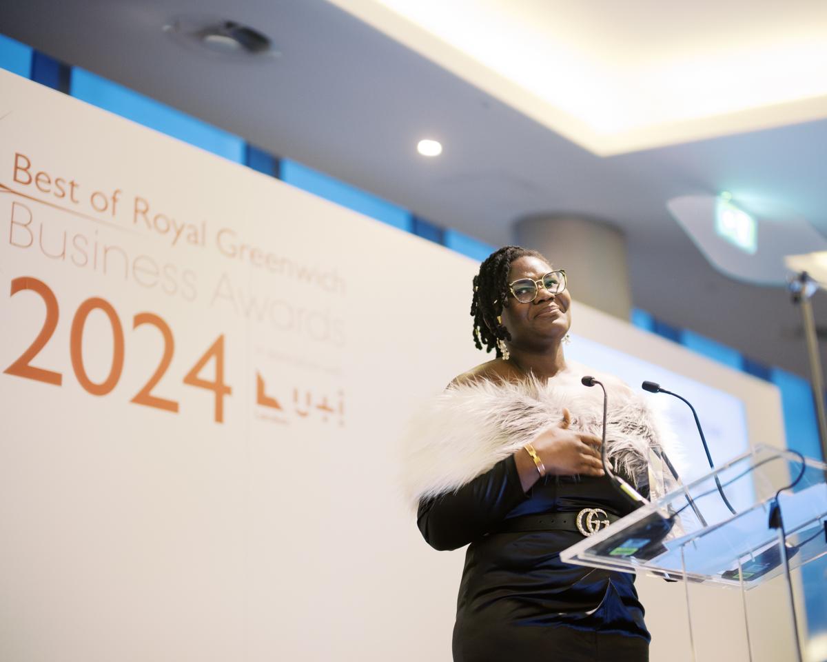 A woman in a black dress and white faux fur stole stands on stage behind a glass lecture, smiling and looking to her right. She has one hand on her chest. Behind her is a white background with red writing reads: Best of Royal Greenwich Business Awards 2024