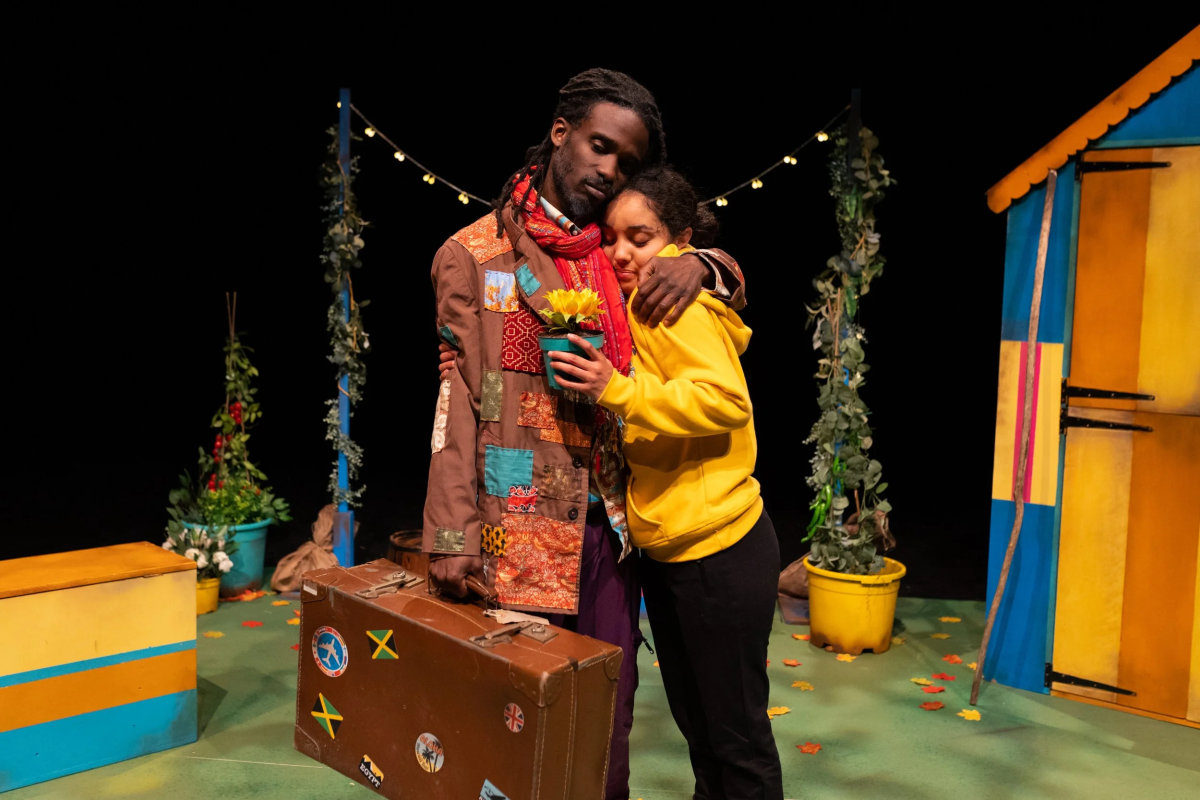 Two people hugging. One is wearing a brown coat and carrying a leather suitcase with Jamaican flags on. The other is wearing a yellow hoodie.