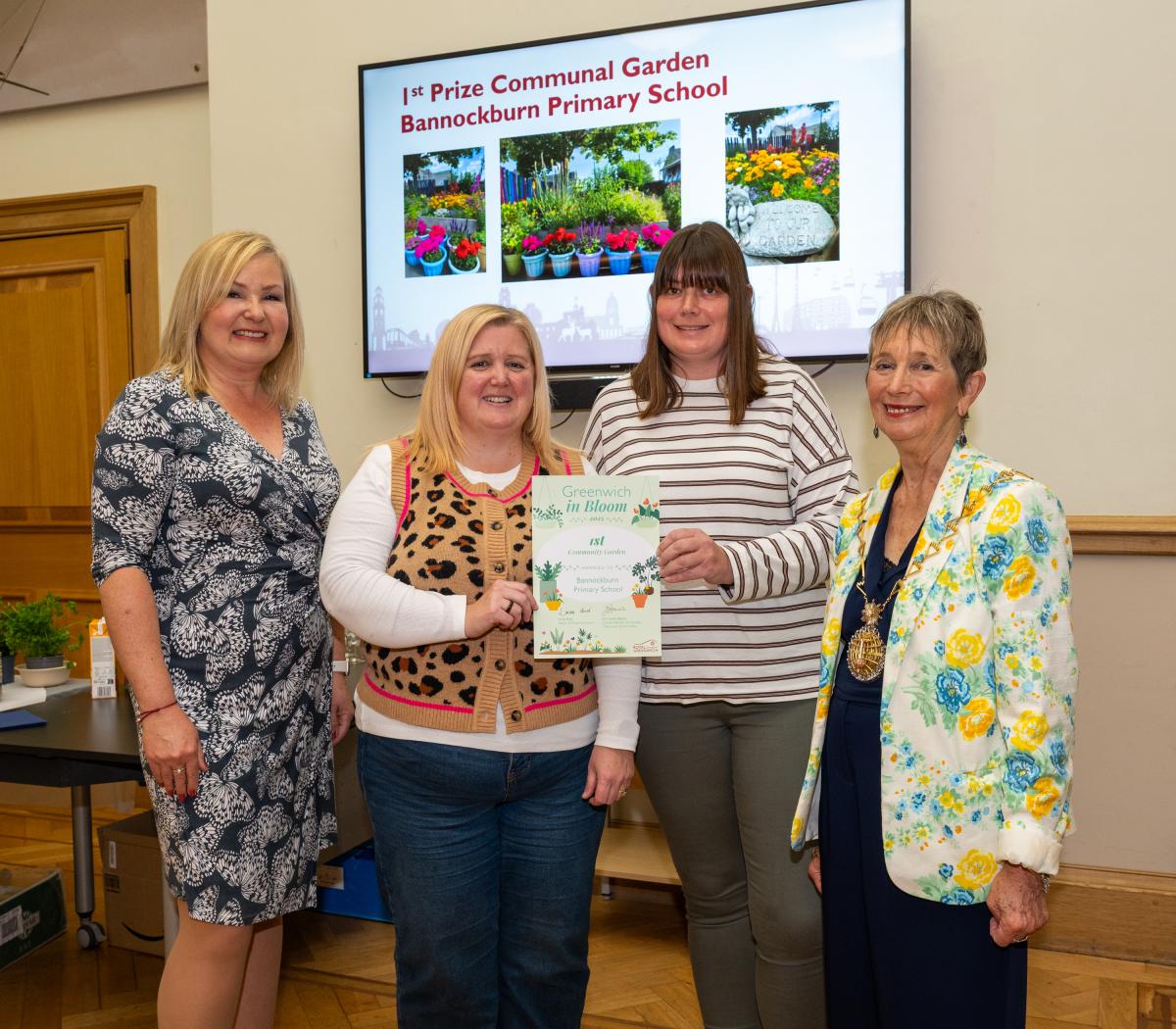 Four people smiling at the camera. The two people in the middle are holding a certificate. Behind them a screen is showing photos of a garden.