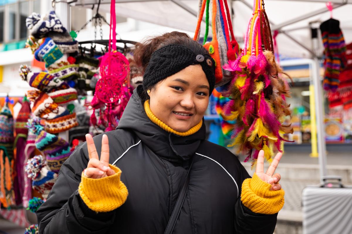 A young woman in a black coat, mustard jumper and black headband stands in front of a colourful market stall. She is giving the peace sign with both hands and smiling to camera.