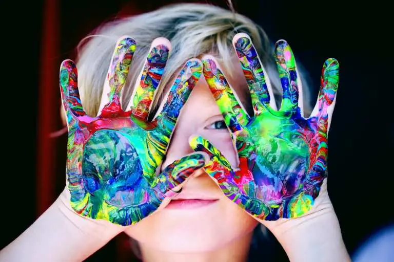 A young boy holding up his hands that are painted in vibrant colours.