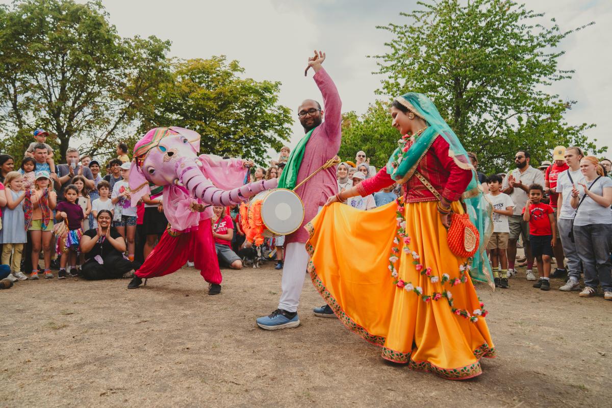 Two performers in colourful clothing. One is dancing and one is drumming, both in front of a crowd. A third dancer is dressed as a pink elephant,