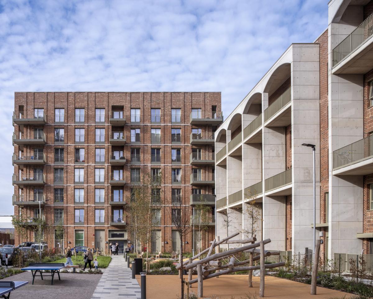 A photo of Kidbrooke Park Road North, the first phase of a 452 home carbon neutral social housing development. The image shows Modern residential buildings with brick and concrete facades surrounding a landscaped courtyard featuring trees, benches, and a wooden climbing structure.