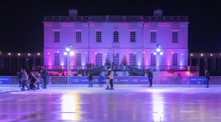 The Queen’s House Ice Rink lit up in purple with several groups of people skating in front of the house.