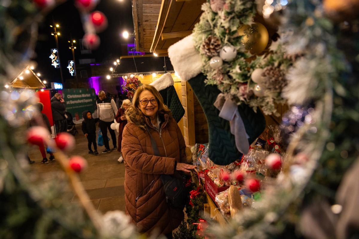 A woman is stood next to a Christmas market stall smiling to camera. There are festive wreaths around the stall.