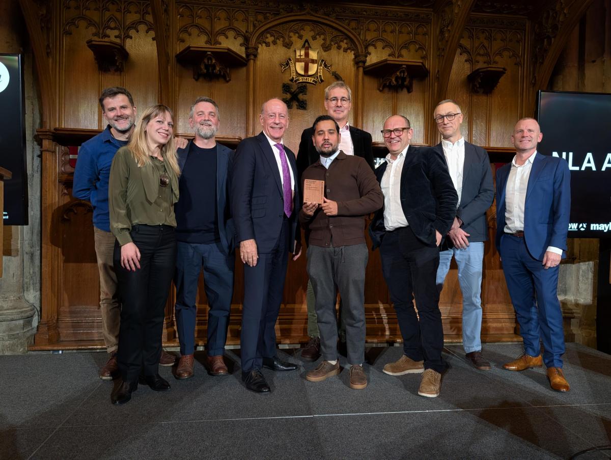 Group of people standing on a stage in a historic hall during the NLA Awards ceremony. Ornate wooden panels and a crest are visible in the background. Councillor Majid Rahman is holding the Mayor’s Prize award plaque while posing with others.