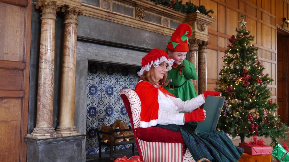A person dressed as Mrs Claus reading a storybook. A person next to her is dressed as an elf.