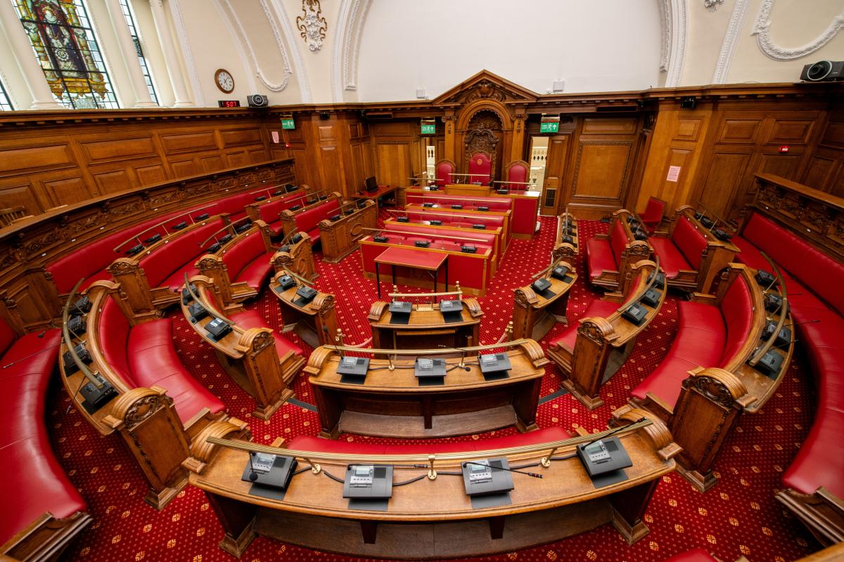 Council chamber room with red carpet and upholstery and wooden panelling. The rows of wooden benches in a semi circle face towards a speaker's chair. The room is empty. 