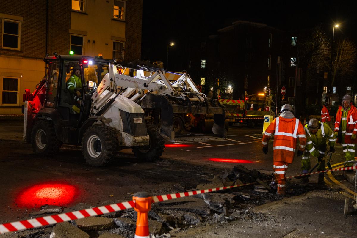 A road being resurfaced at night by a white digger, surrounded by people in orange hi-vis gear.