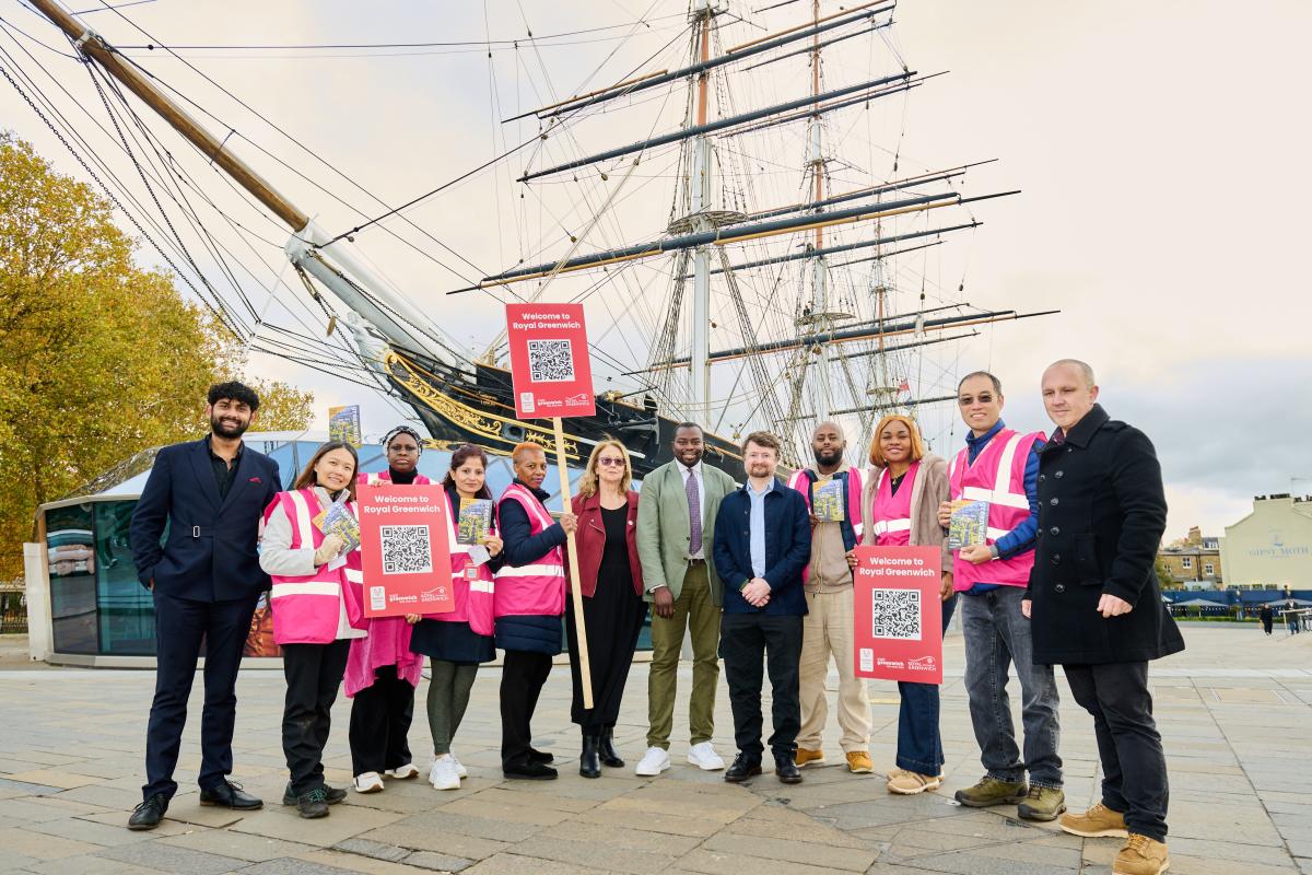 A group of people stand in front of the Cutty Sark. Some are wearing pink hi-vis vests and carrying placards with a QR code