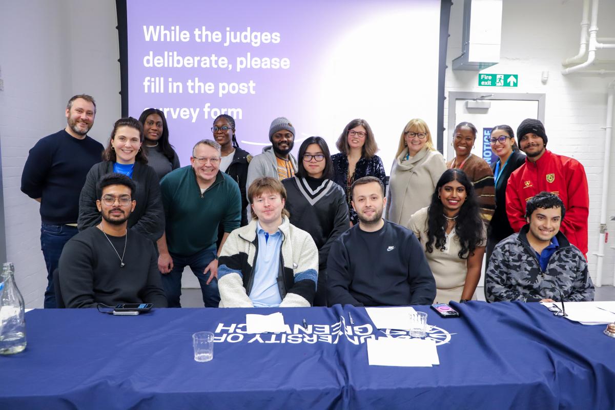 A group of people pose for a photo in front of a table that has a blue table cloth with the University of Greenwich logo. 