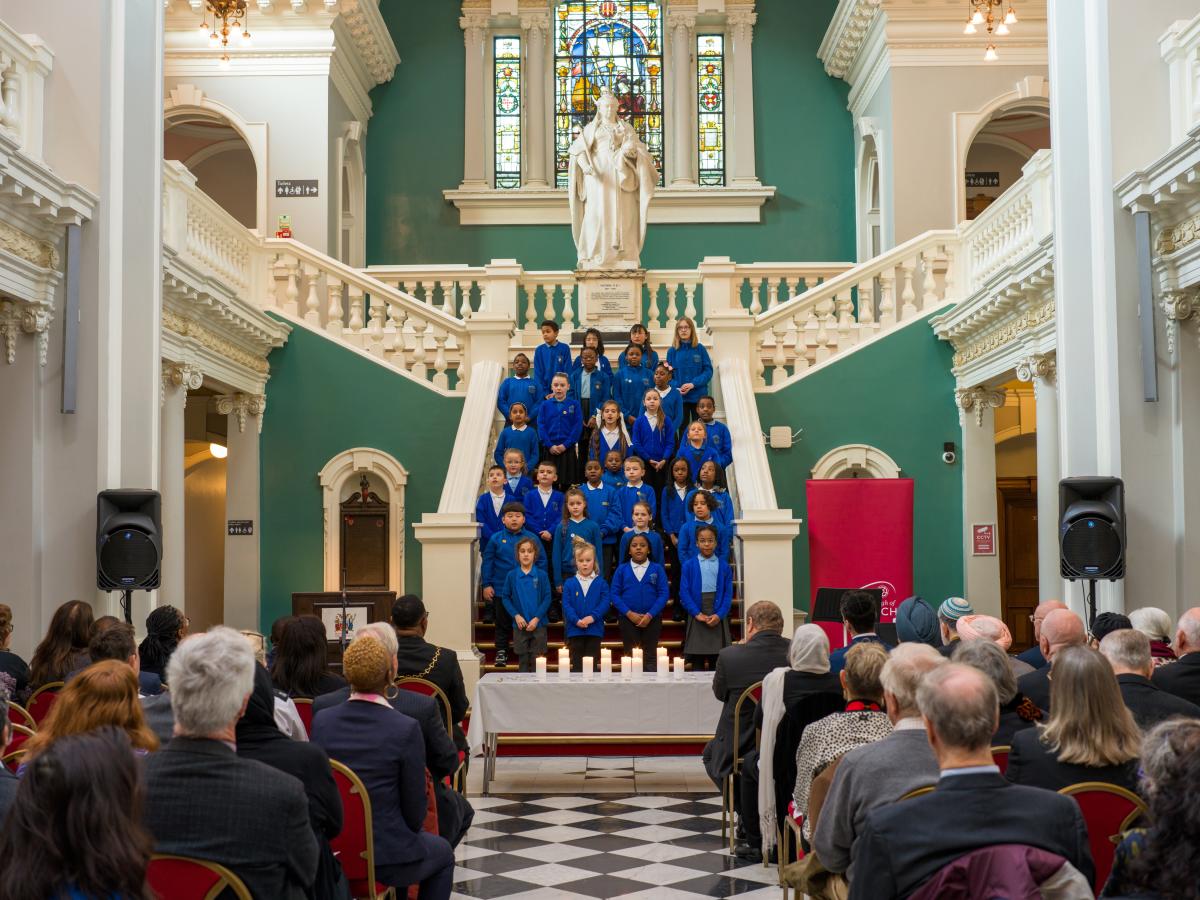 A choir of children wearing blue uniform singing on a large white staircase. In front of them is an audience of people with their backs facing the camera.