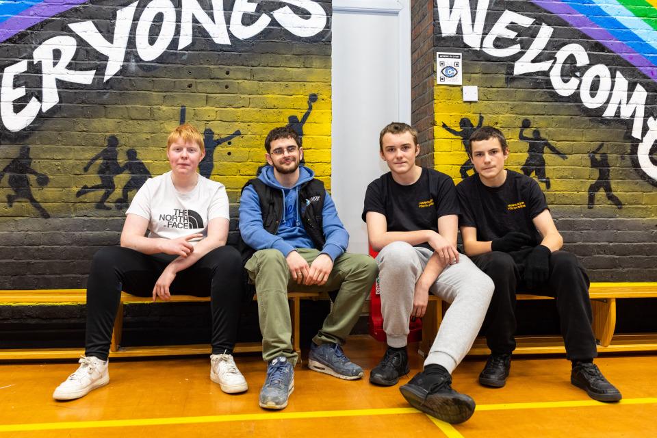 Four young people at a youth club sitting in front of wall art that reads ' everyoe's welcome'
