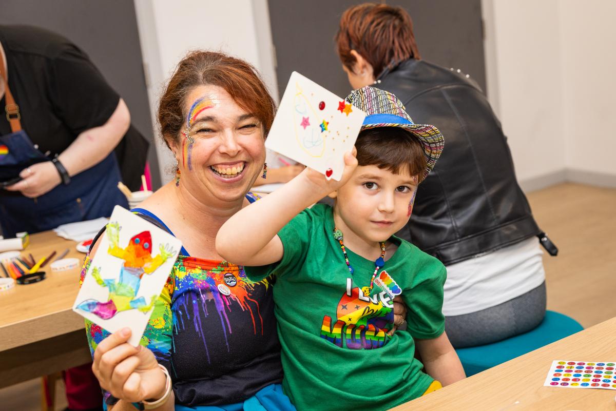 An adult and a child sat at a table. They are holding up rainbow coloured artworks.