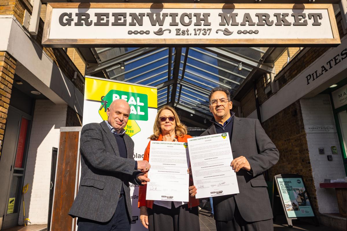 Three people stand outside the entrance of Greenwich Market holding a charter. Behind them is a pull-up banner that reads: Real Deal