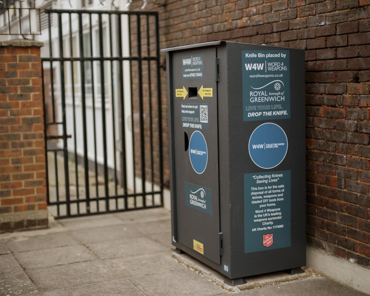 Grey weapon surrender bin in Abbey Wood, placed against a brick wall and fenced area.