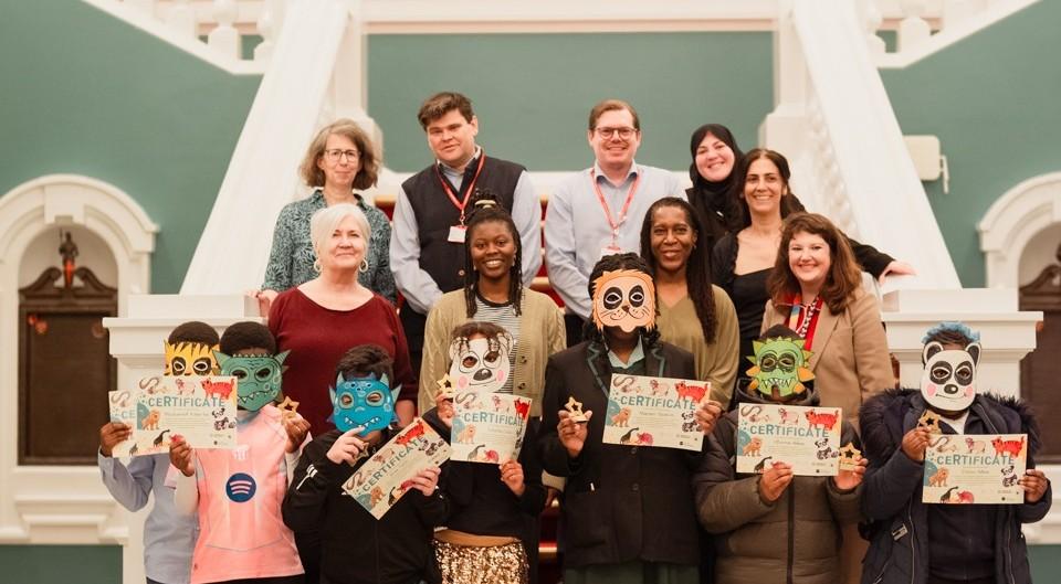 Children wearing masks and holding their work, posing for a photo in the Town Hall with council officers.