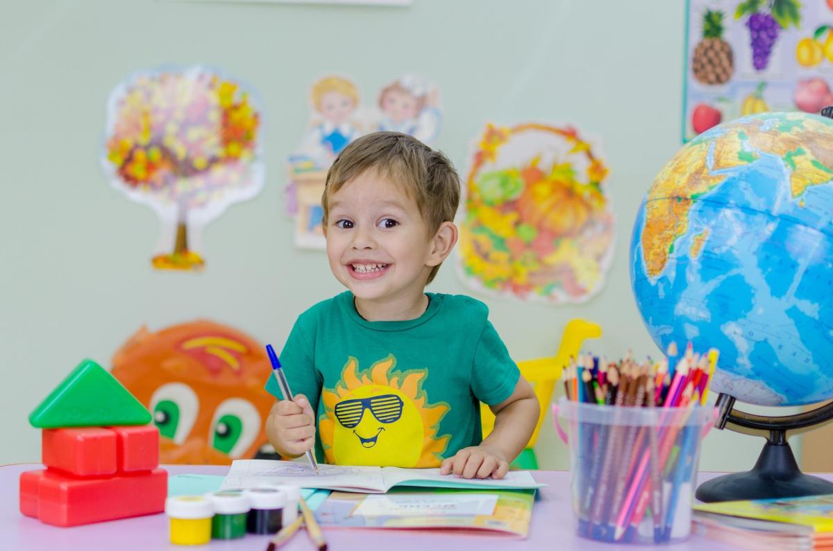 Child at school smiling to camera while he colours in, with a world globe and colouring pencils on table infront of him